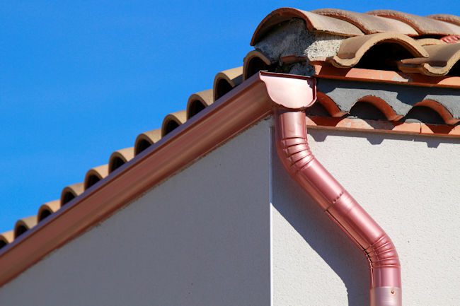 Close-up of a red metal gutter and downspout installed on a tiled roof with clear blue sky, showing professional roof and gutter installation