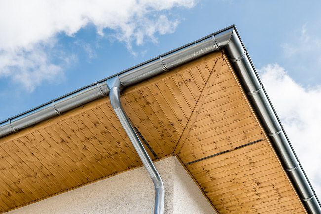 Close-up of residential roof soffit and seamless gutter installation with metal downspout against blue sky