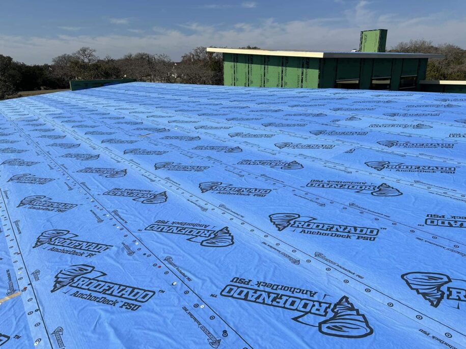Blue weather-resistant roofing underlayment with tornado branding installed on a flat commercial roof during construction, with green building structure visible in background
