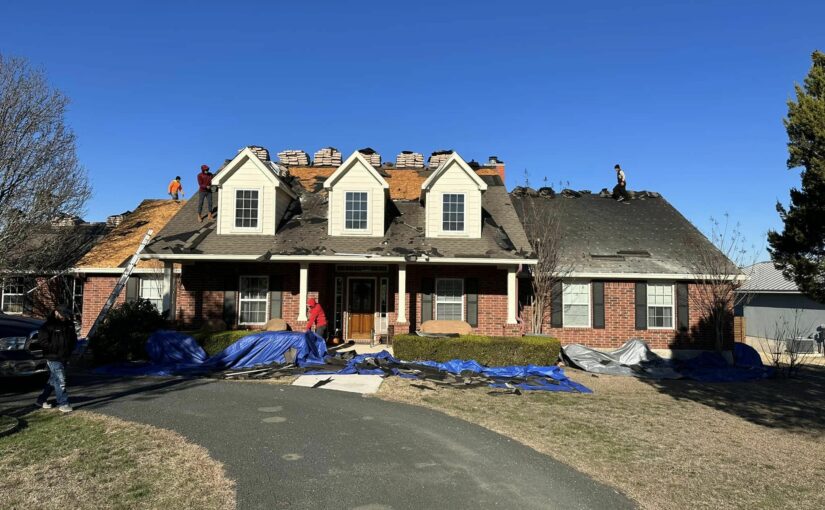 Brick residential home undergoing roof replacement with workers on roof and blue tarps covering yard in San Antonio