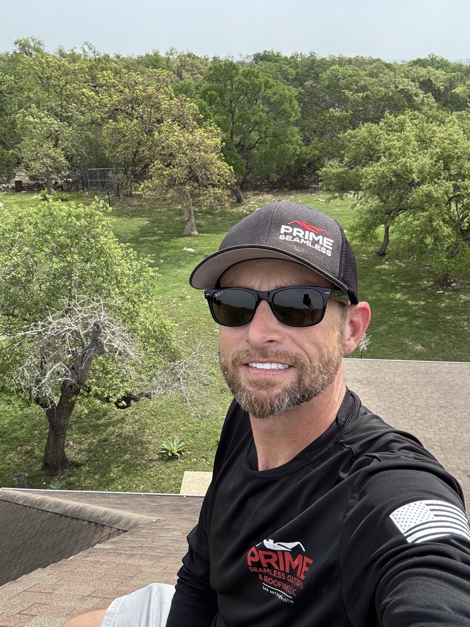 Man in Prime Seamless Gutters and Roofing branded apparel smiling at camera with lush green orchard landscape behind him in San Antonio, Texas