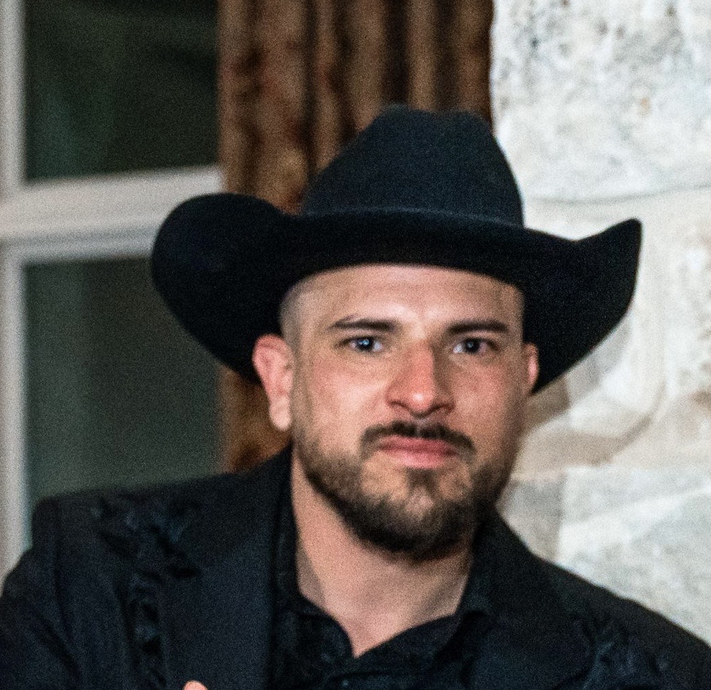 Man wearing black cowboy hat and black shirt with goatee, indoors with blurred background