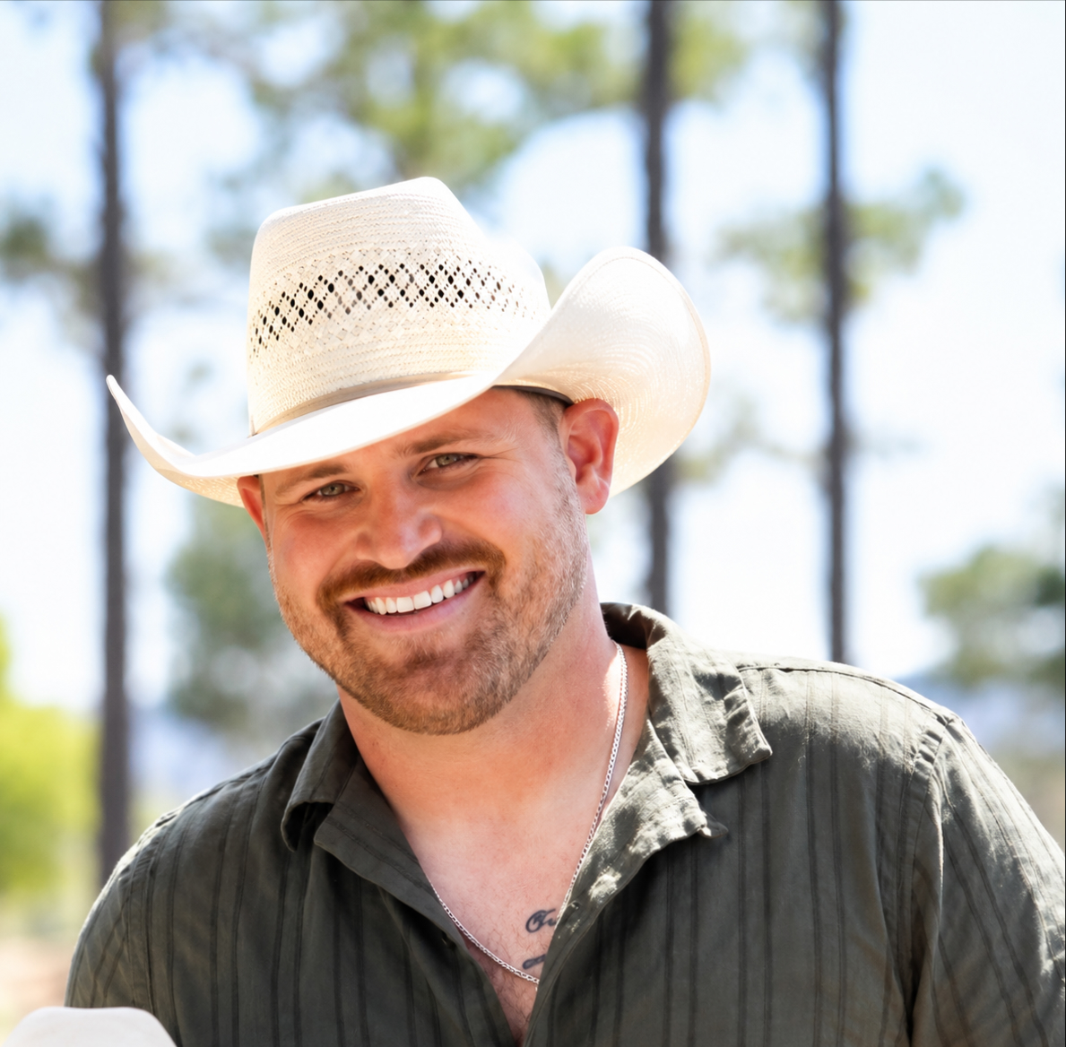 Smiling man wearing a cream cowboy hat and dark striped shirt outdoors with trees in background