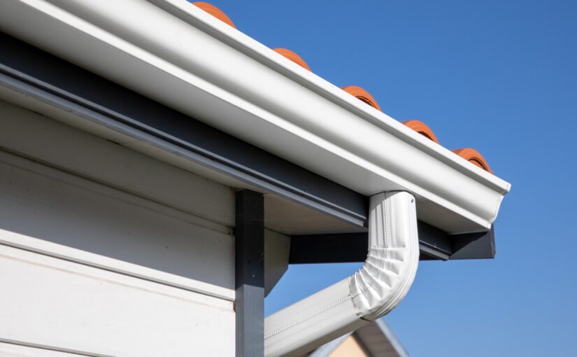 White seamless gutters and downspout installed on residential roofline with orange roof vents against clear blue sky