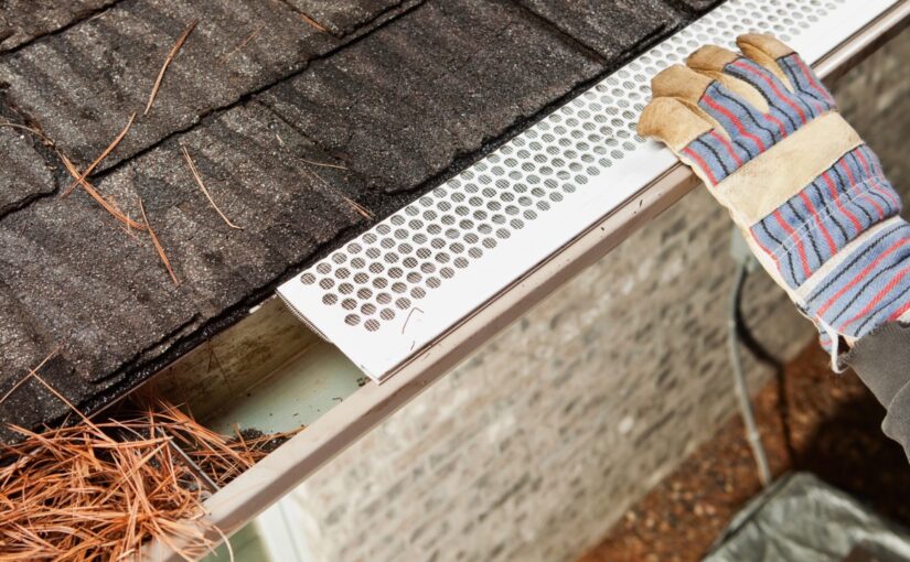 Worker in striped gloves inspecting white gutter guard on roof edge with shingles and pine needles visible, gutter maintenance and roof protection